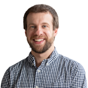 White man with light brown hair and facial hair wearing a blue and white plaid button down shirt.