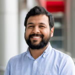 Photo of an asian man with short black hair and beard who is smiling and wearing a light blue button down shirt