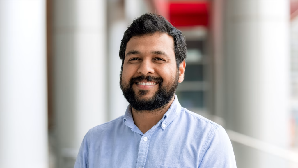 Photo of an asian man with short black hair and beard who is smiling and wearing a light blue button down shirt